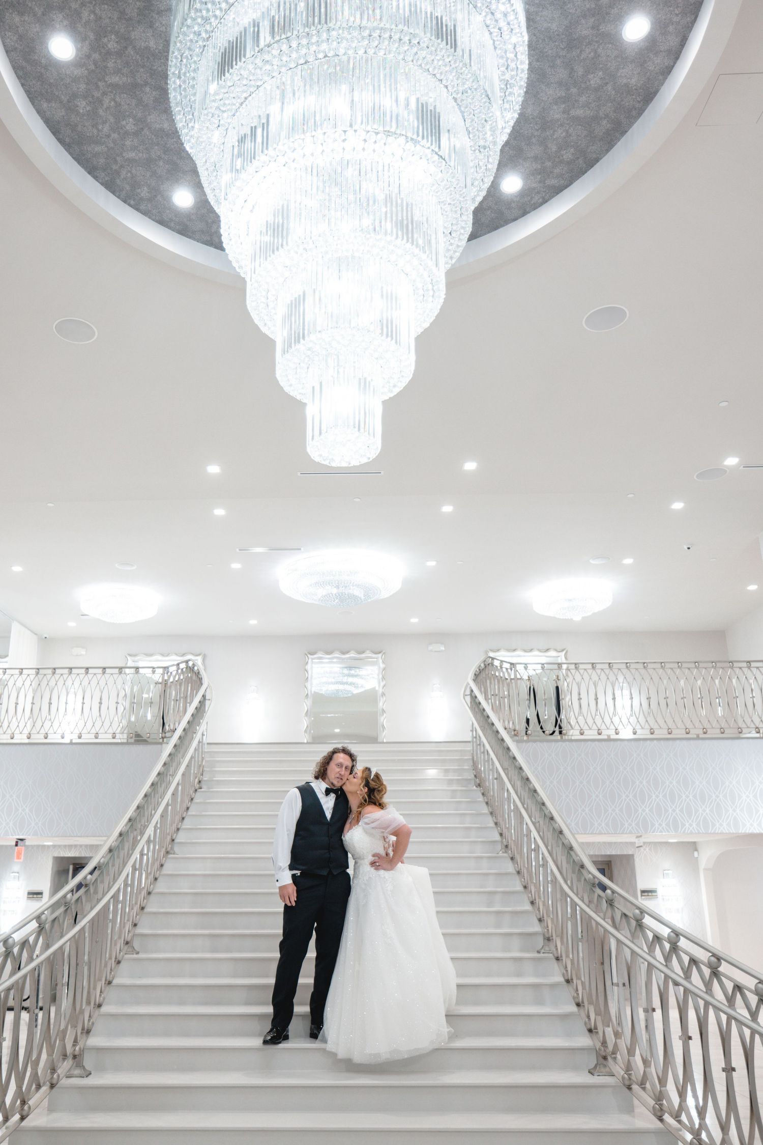 Elegant white staircase with crystal chandeliers in a wedding venue showcasing ornate railings and bright lighting.