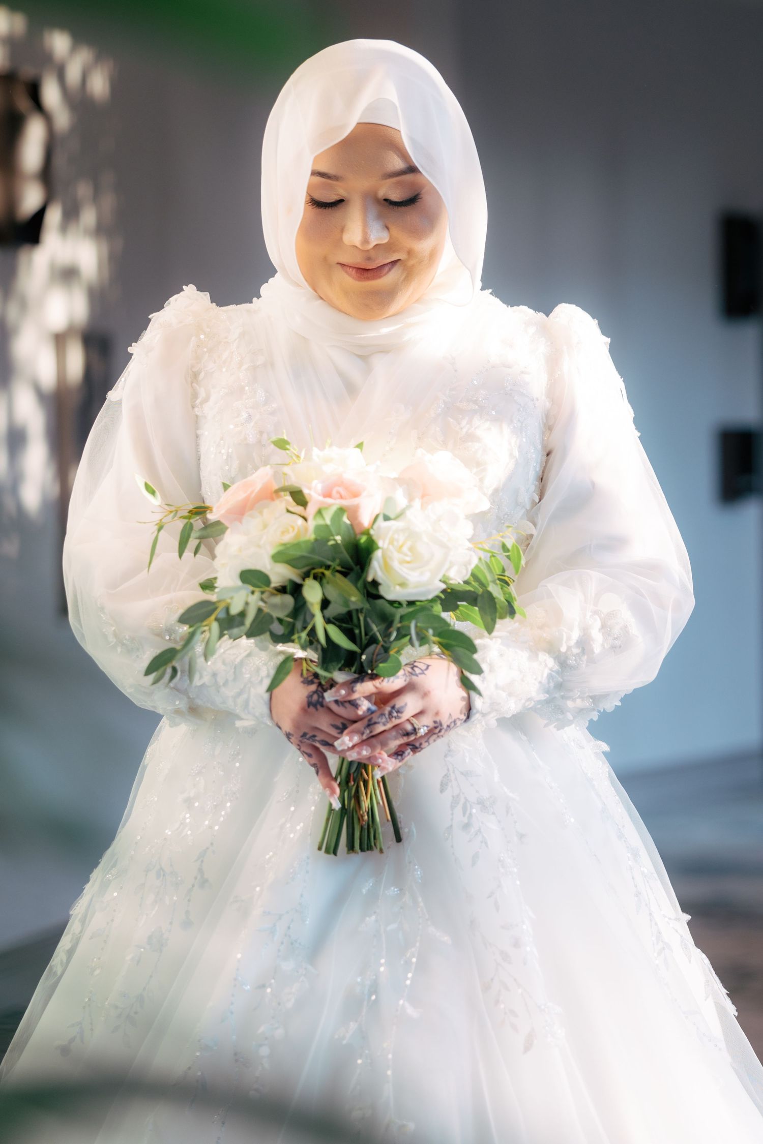 A bride in a white hijab and elegant wedding dress holds a delicate bouquet of flowers and greenery.
