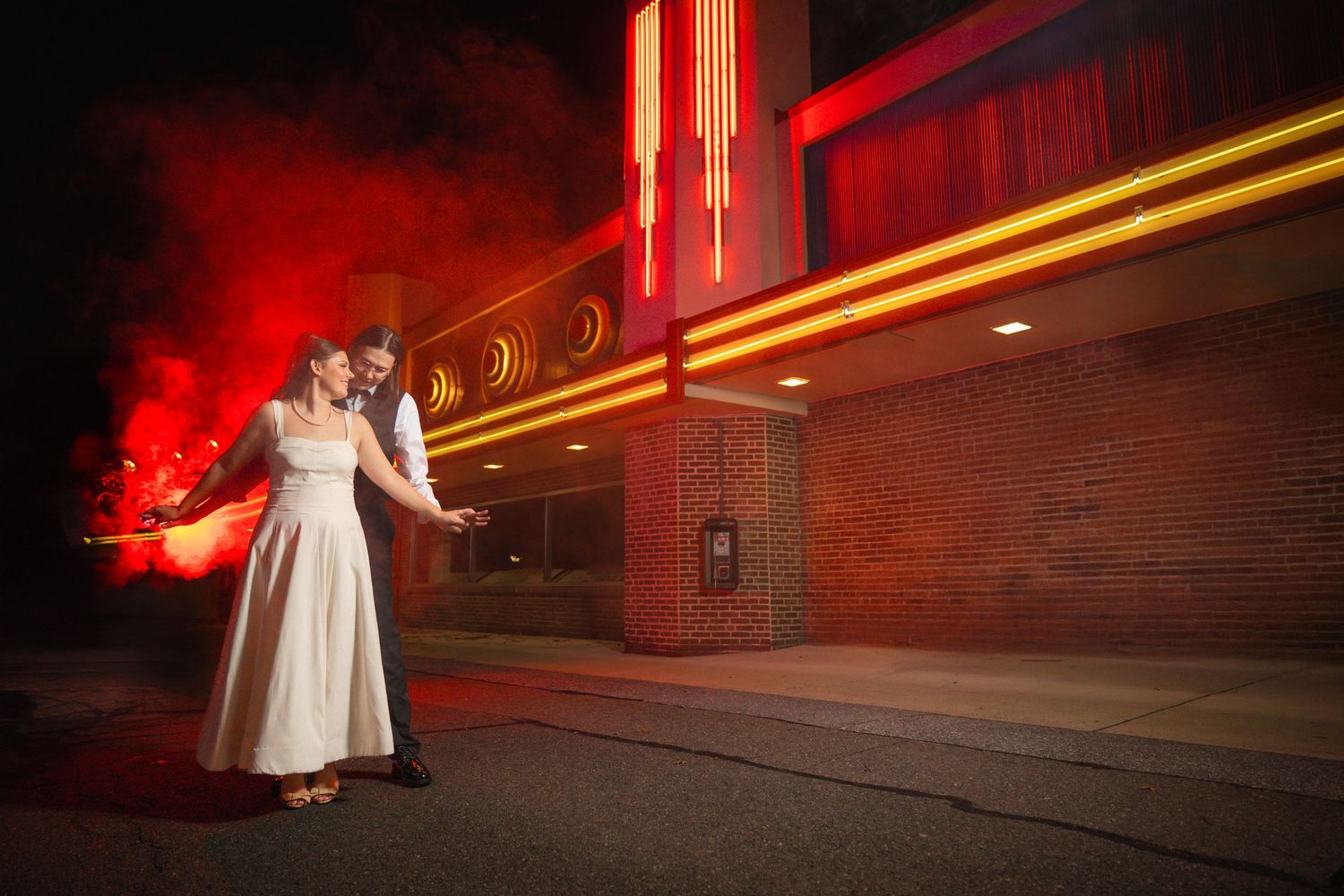 A couple in wedding attire dances together under dramatic red lighting at an outdoor venue during nighttime.