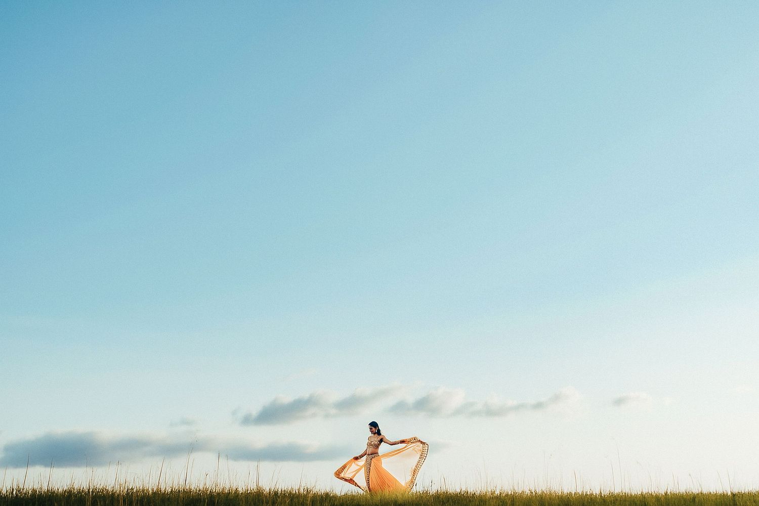 A silhouette against a blue sky stands in tall grass holding a flowing fabric that catches the wind.