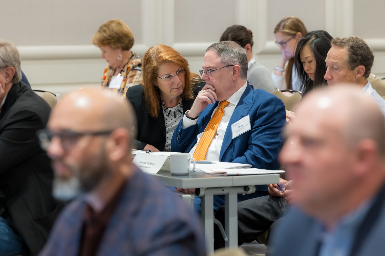 Business professionals sit attentively in rows during a conference or training session in a bright meeting room.