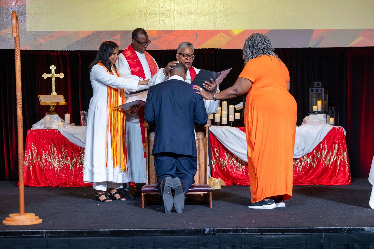 A series of people in religious robes and ceremonial attire gather on a dark stage during a church service.