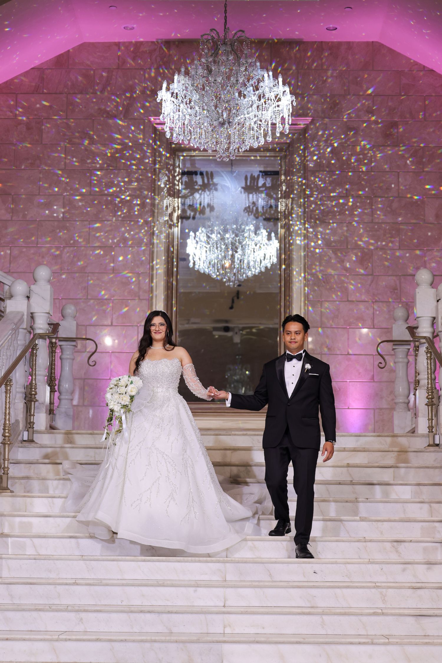 A wedding couple descends grand white marble stairs under a crystal chandelier with pink lighting in an elegant venue.
