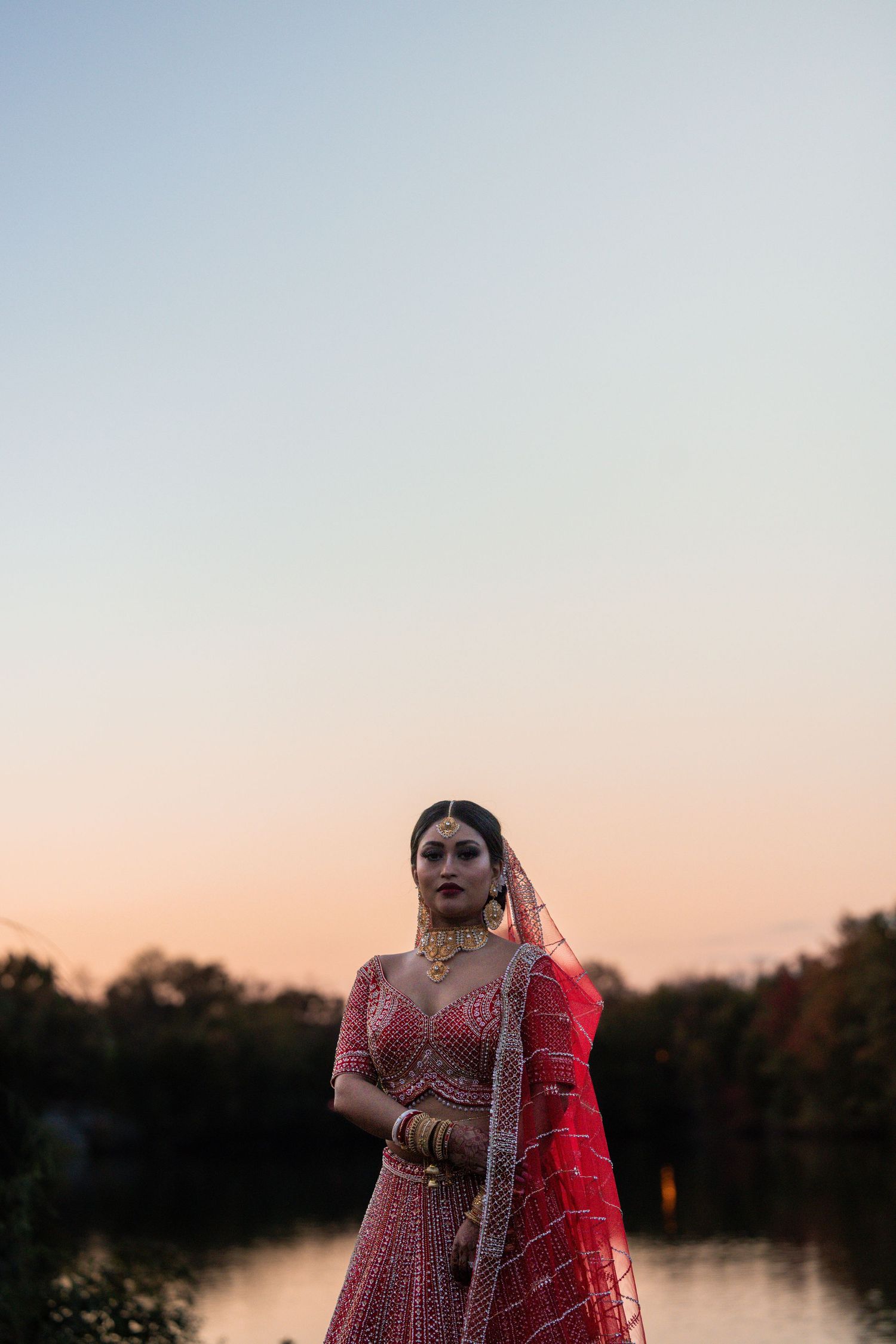 A bride in a traditional red and gold Indian wedding dress poses gracefully by a serene lake at sunset.