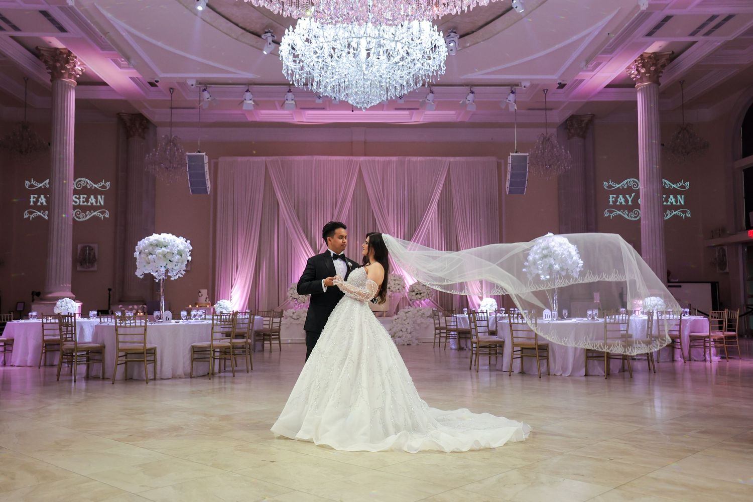 Elegant ballroom decorated in soft pink lighting with crystal chandeliers and white tables for a luxurious wedding reception.