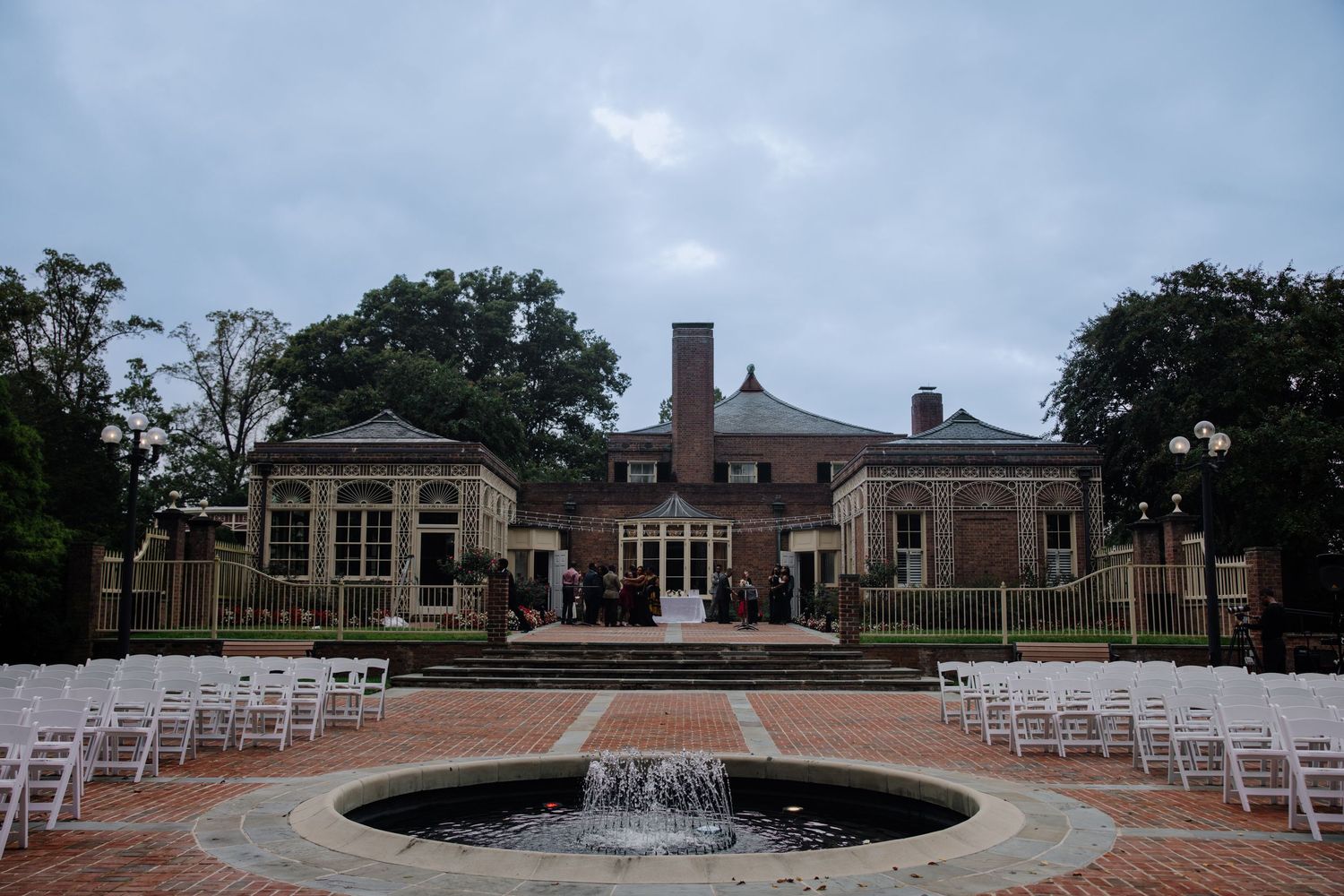 A panoramic view of an outdoor wedding venue with white chairs set up on a brick patio near a circular fountain.