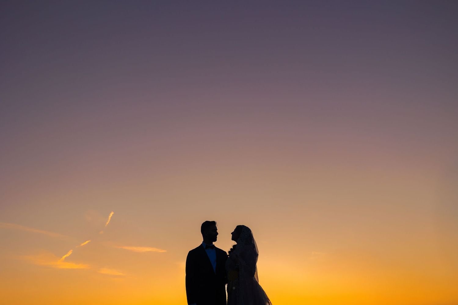 Two silhouetted figures stand close together against a vibrant orange and purple sunset sky.