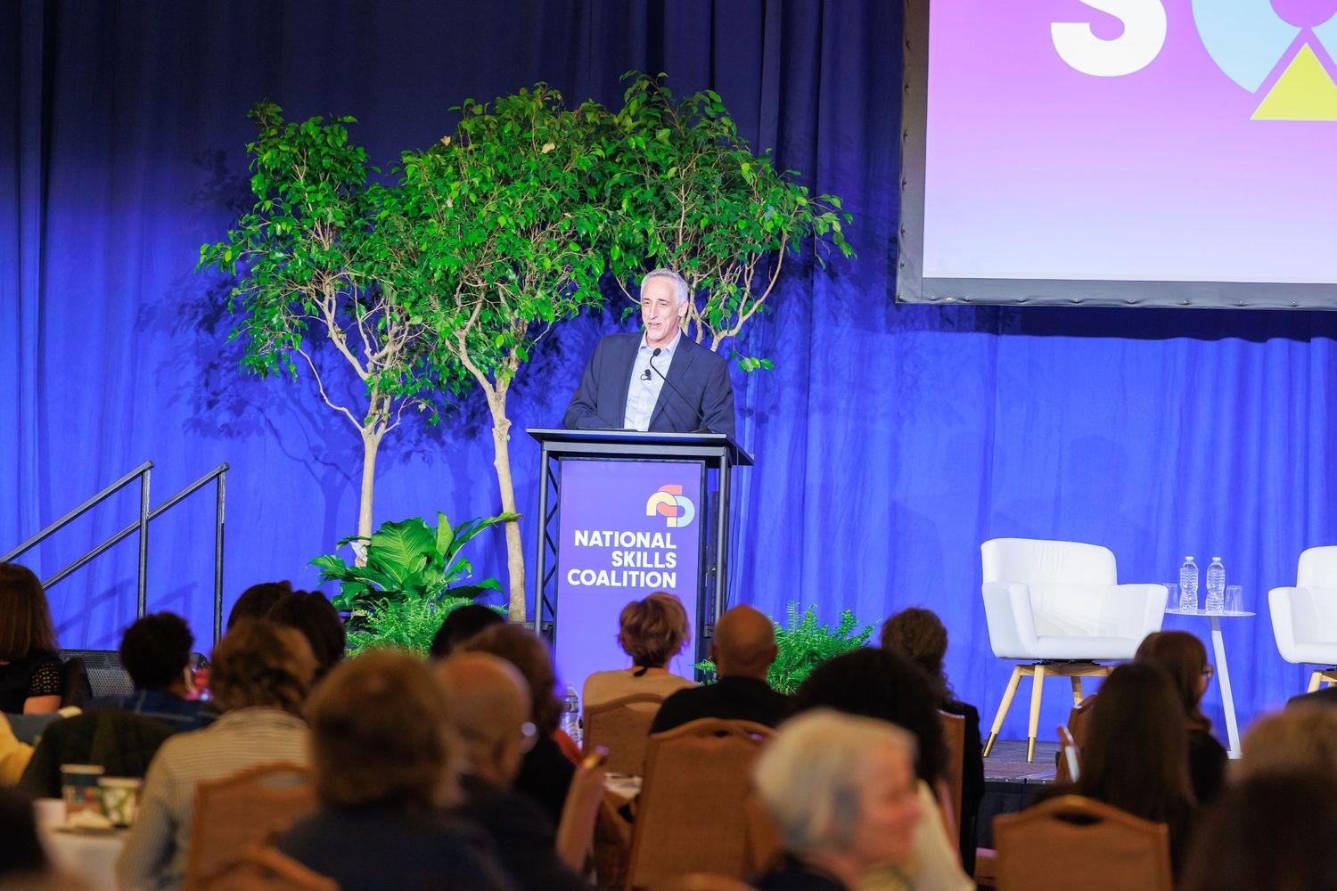 A large audience sits in a conference hall with blue lighting and stage decorations featuring green plants.