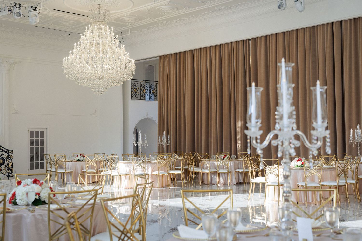An elegant banquet hall with crystal chandeliers, white tables, and brown curtains set for a formal event.