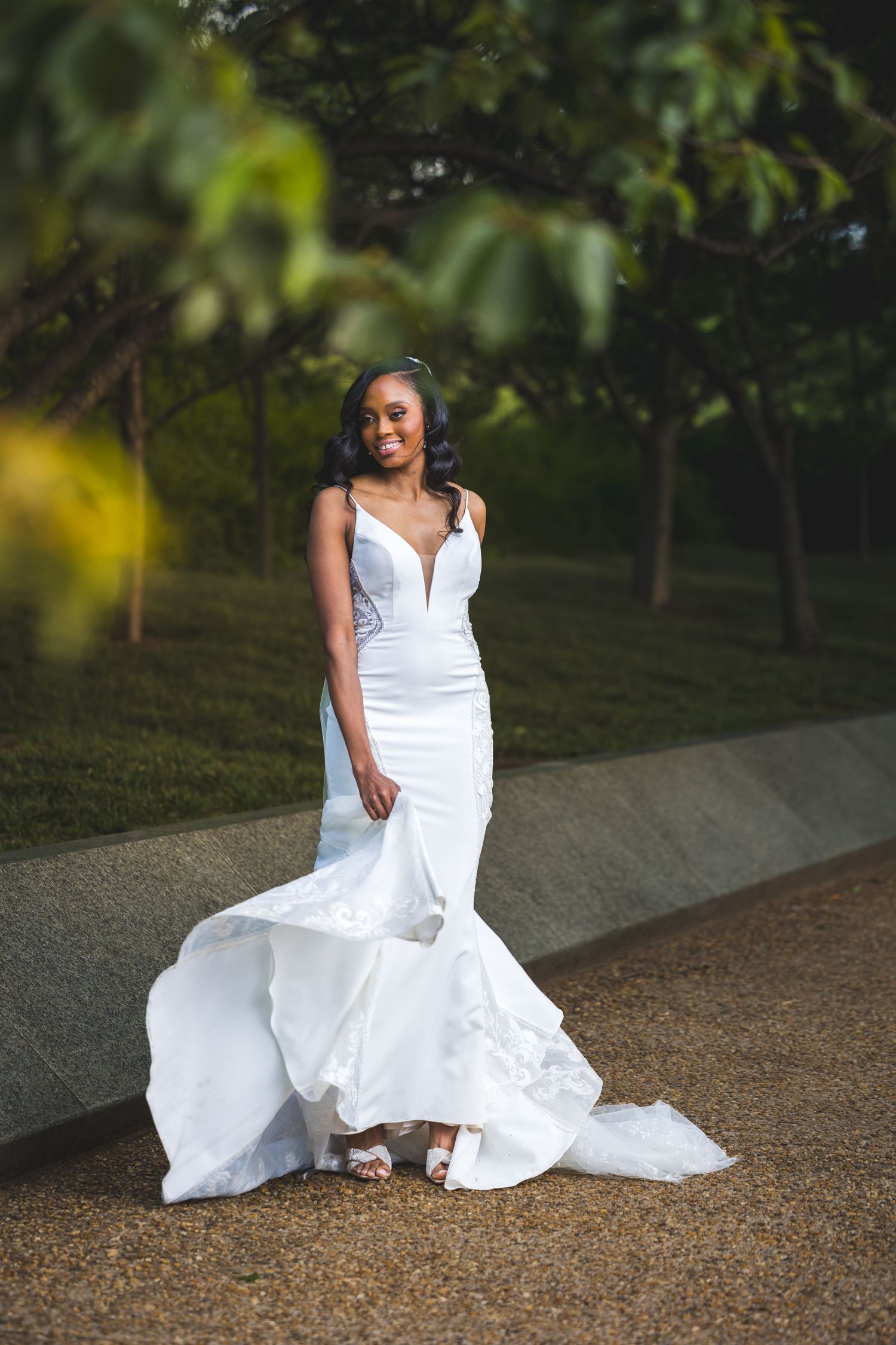 An elegant white mermaid wedding gown with plunging neckline and flowing train on a path surrounded by greenery.