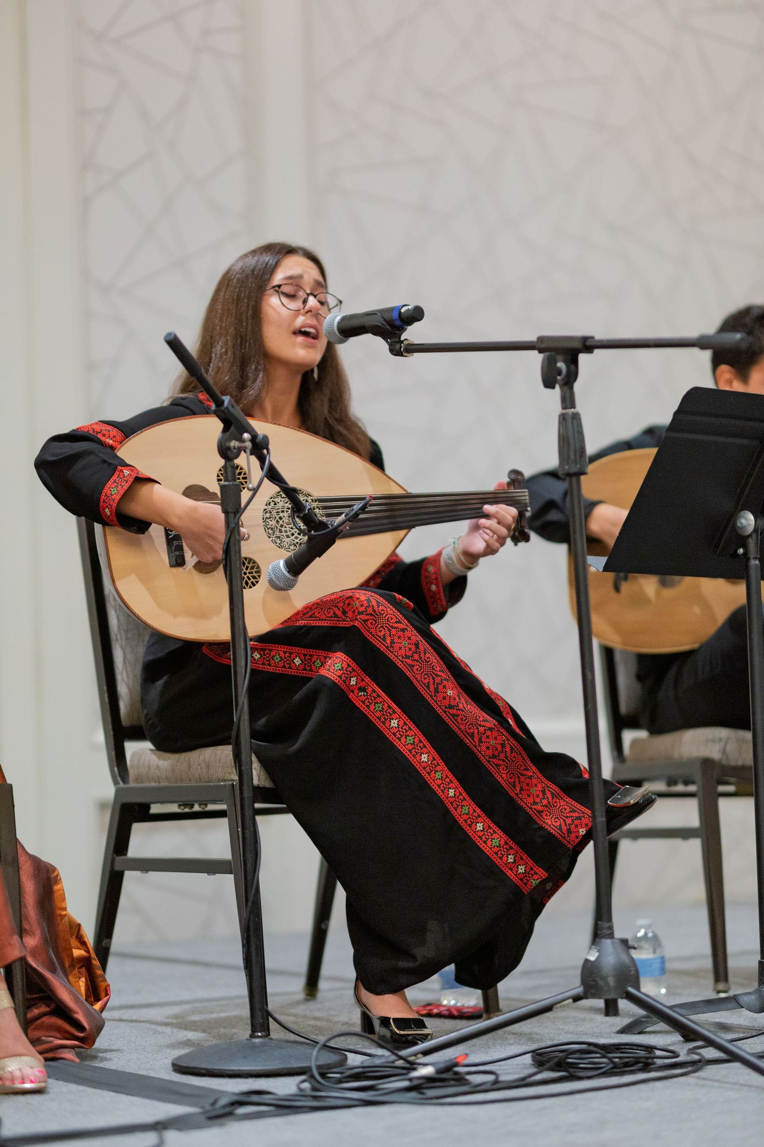 A performer in a black dress with red trim sings and plays guitar while seated on stage during a musical performance.
