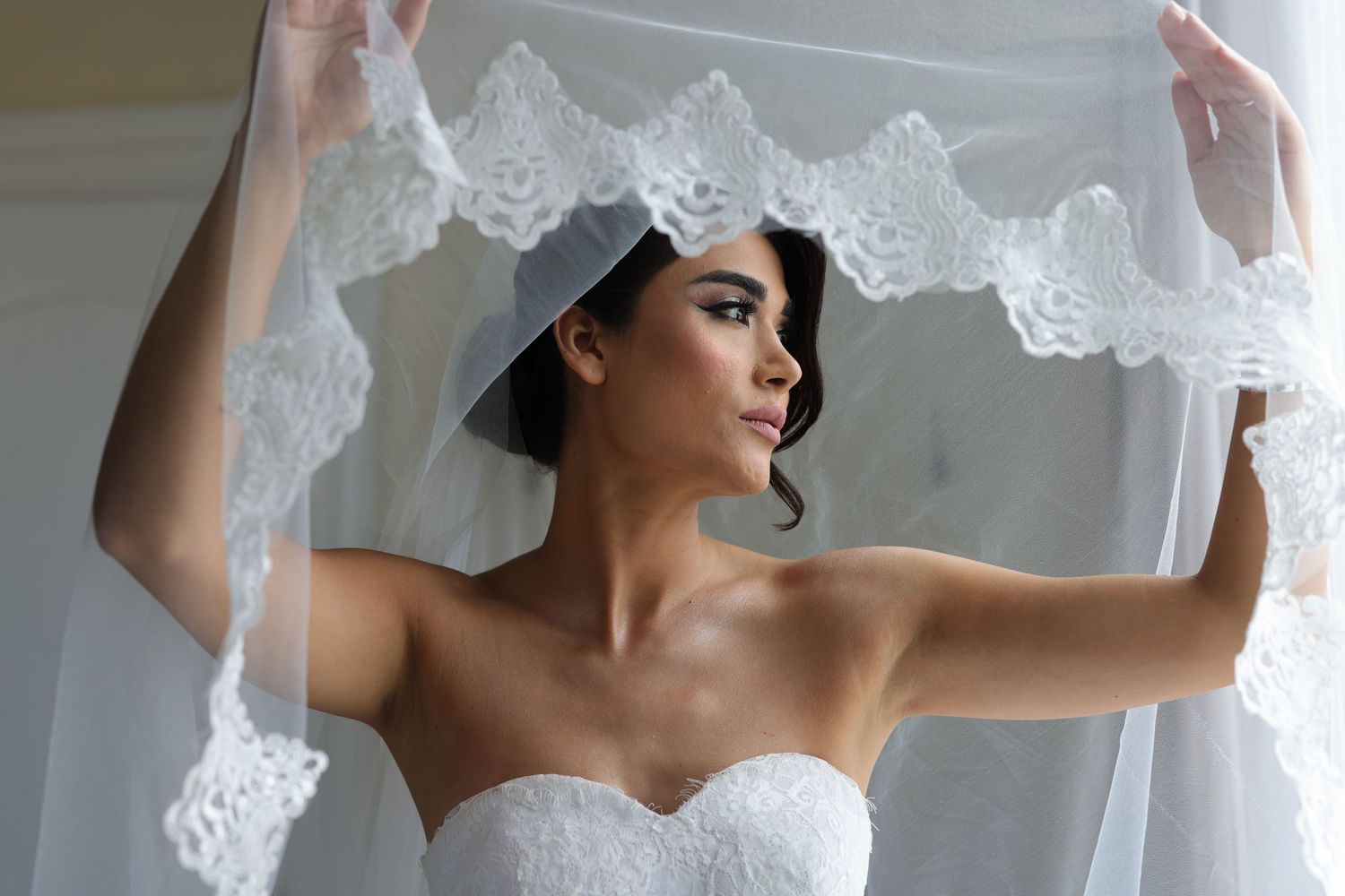 A bride holds up a delicate white lace veil against a light background, showing the intricate details of the bridal accessory.