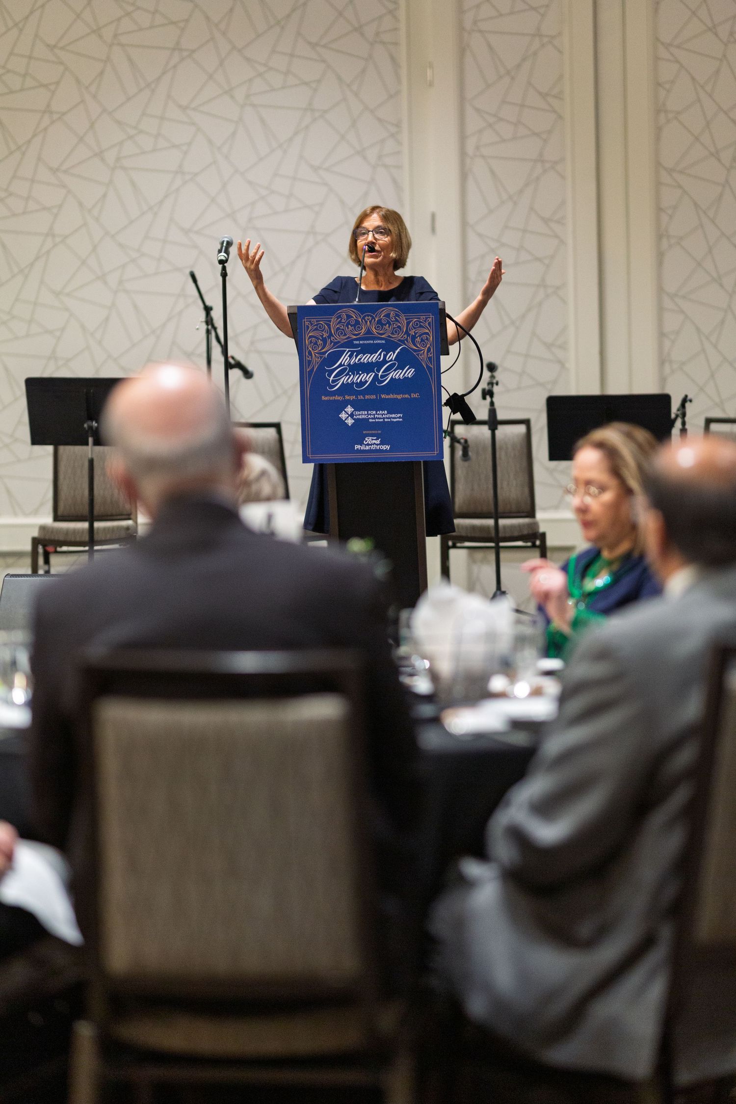 A presenter stands at a podium addressing an audience seated in dark chairs during a business conference.