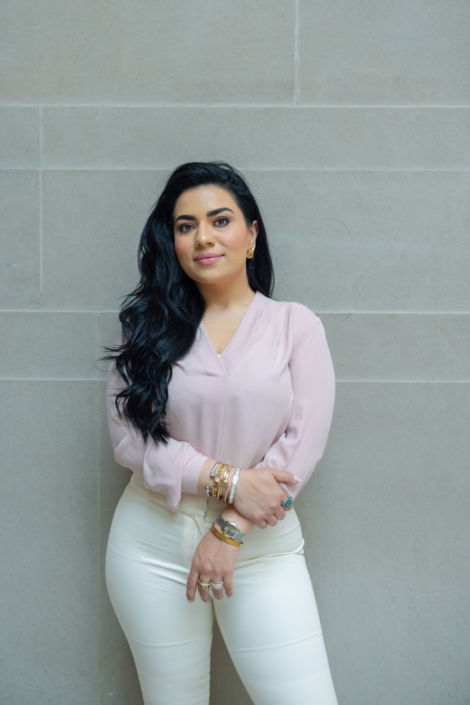 A woman in a pale pink blouse and white pants poses against a grey stone wall while wearing gold jewelry.