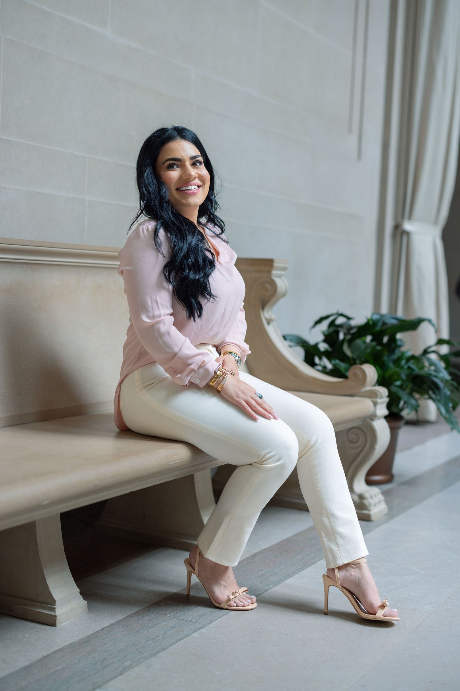 A person in a pink blouse and white pants sits on an elegant stone bench with plants visible in the background.