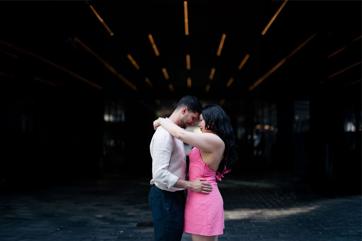 A couple in formal attire embraces in a dark rustic barn with dramatic lighting and beams overhead.