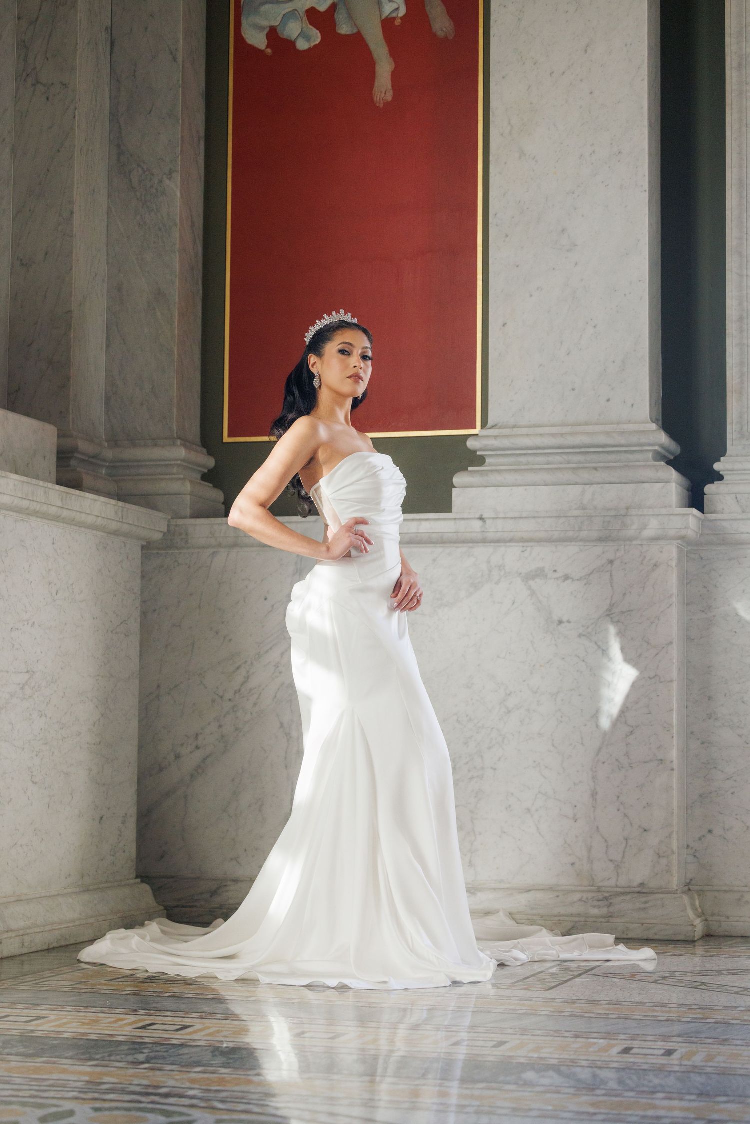 Elegant bride in a white strapless mermaid gown and tiara poses gracefully between marble columns in a grand hall.