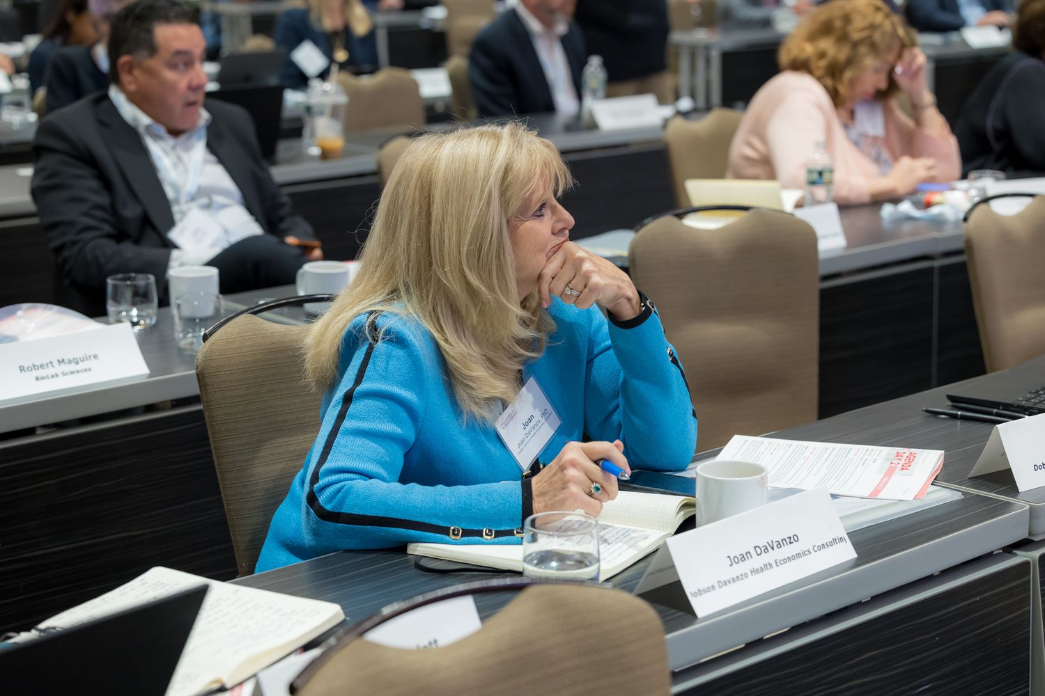 A participant in a blue sweater listens attentively during a professional conference session at a meeting table.