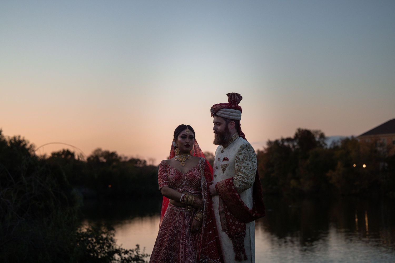 A couple in traditional Indian wedding attire poses against a sunset backdrop near a serene lake.
