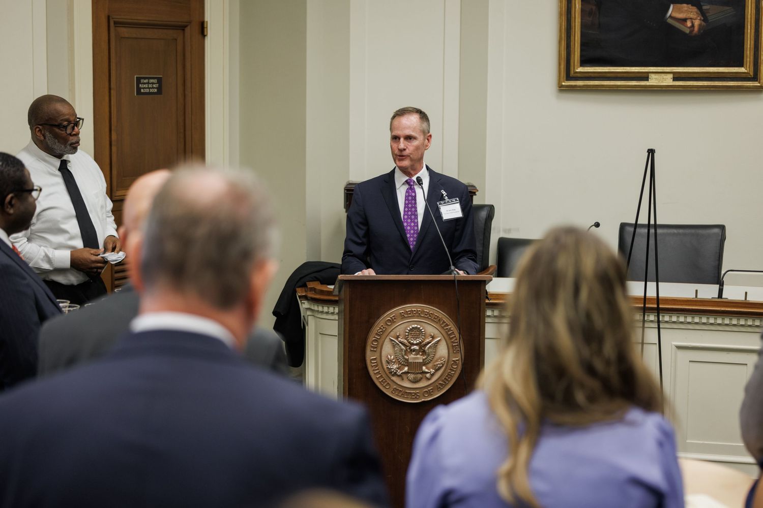 Official speaking at a podium with the U.S. seal in a formal government hearing room with seated attendees.