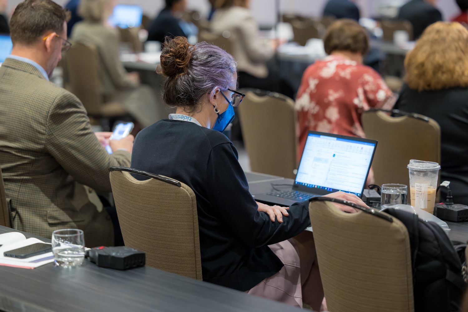 People sit at tables working on laptops during a business conference or training session in a large meeting room.