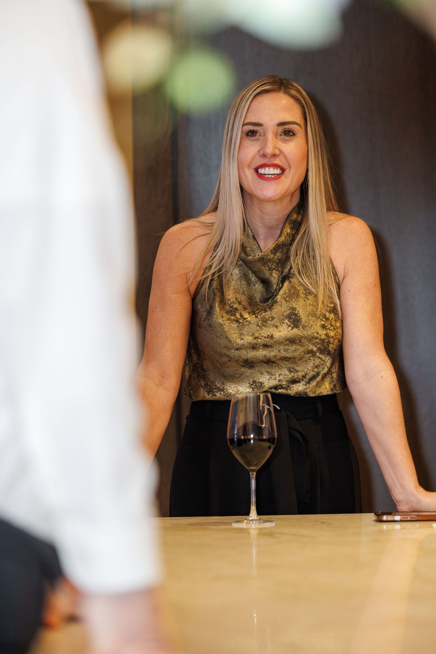 Person wearing a gold sequined top sits at a bar counter with wine glasses in an elegant setting.