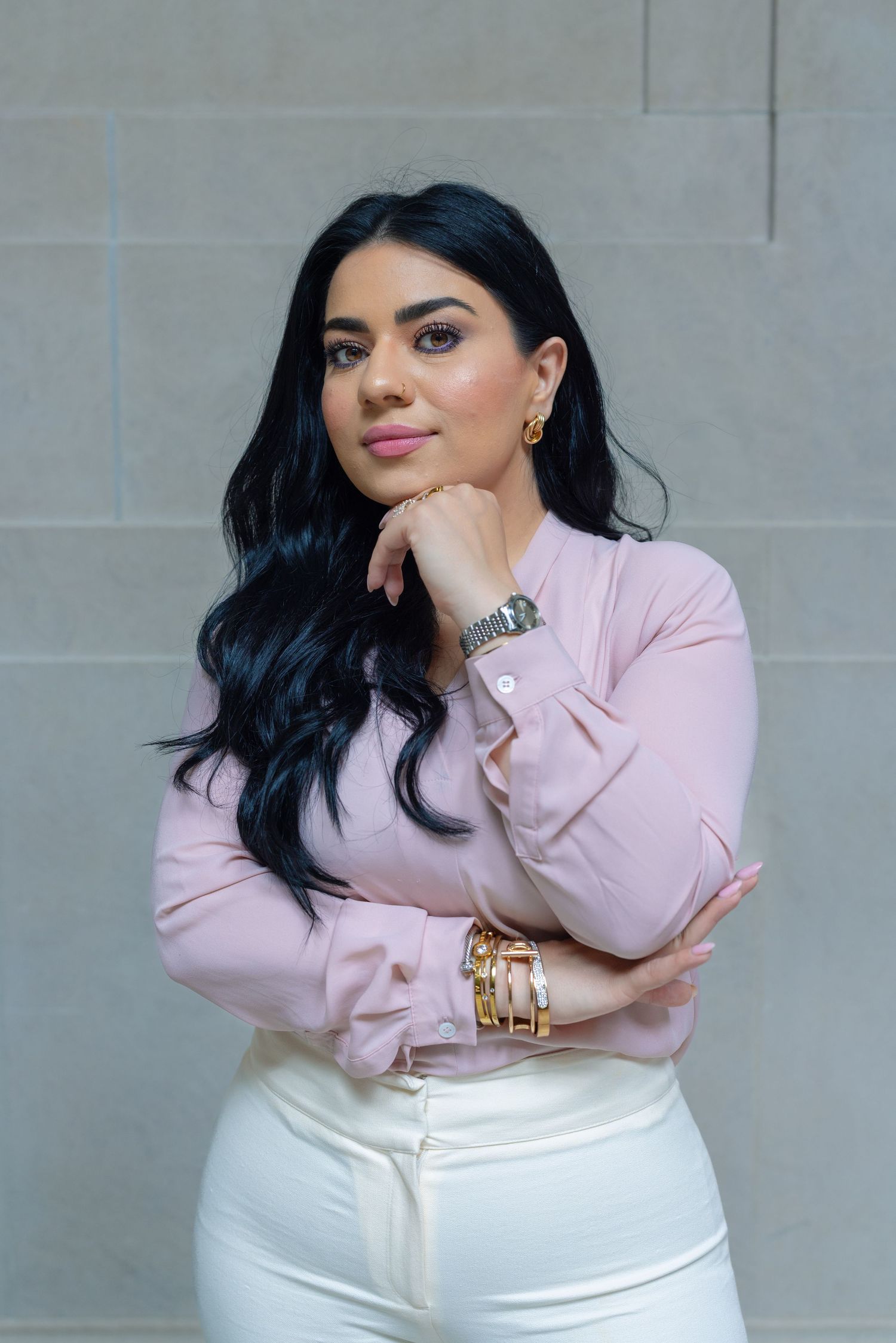 A fashionable woman in a pink blouse and white pants poses against a gray stone wall.