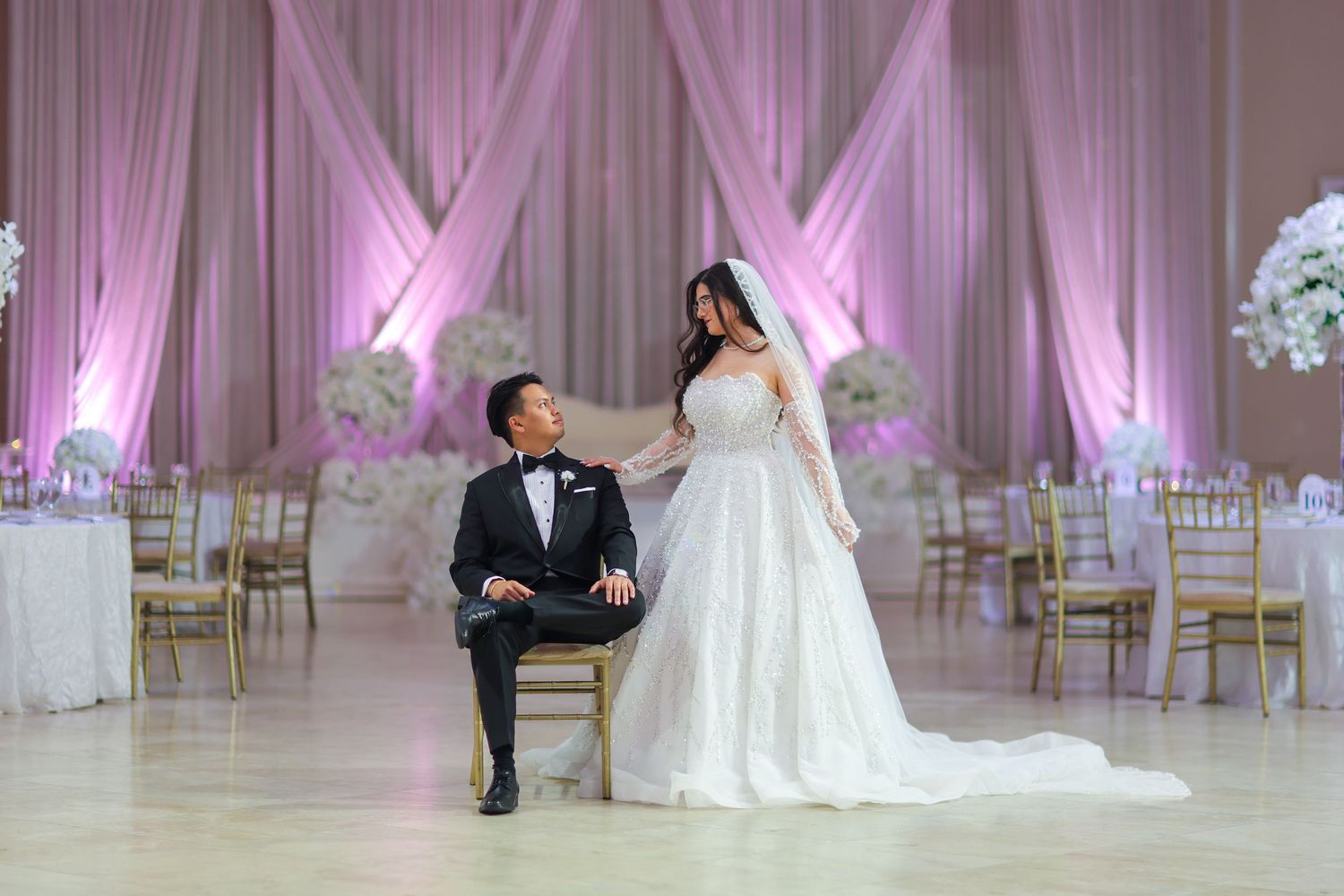 Elegant wedding reception venue with pink uplighting, white drapes, and a couple posing in formal attire.