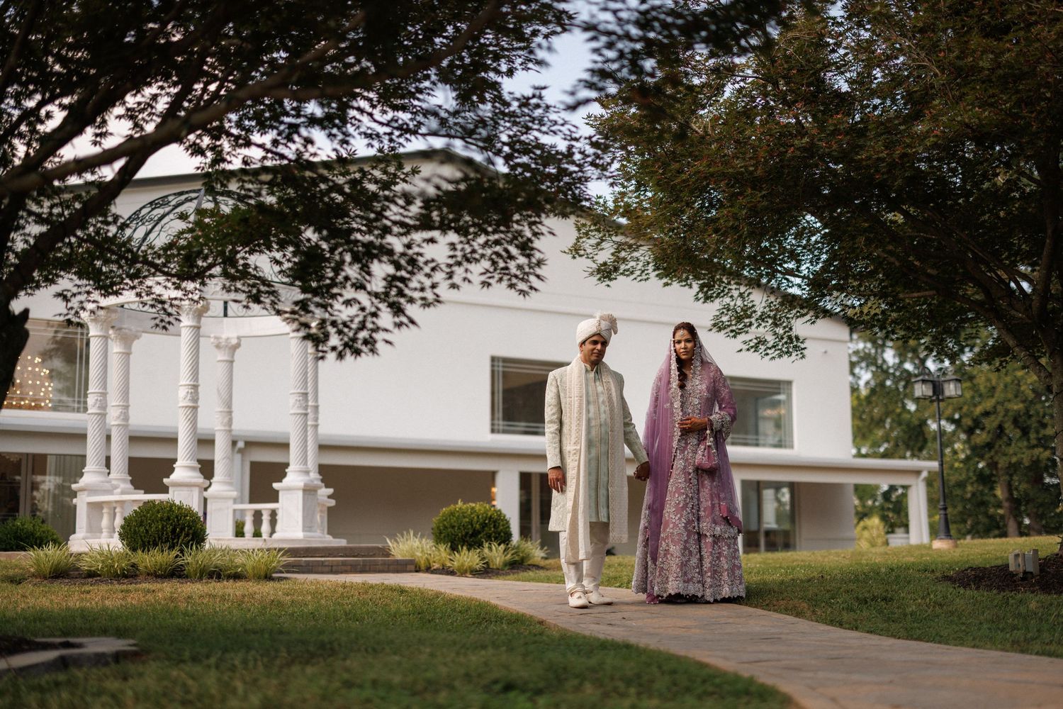 Couple walking together on a paved path leading to a white house with trees and landscaping in the background.