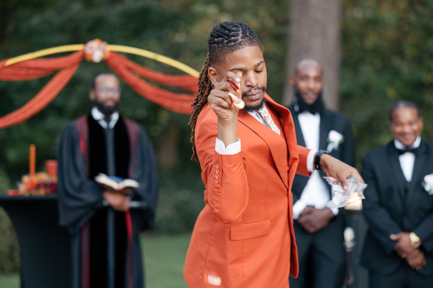 Someone in a bright orange outfit expresses joy during an outdoor wedding ceremony with officiants in black robes in background.