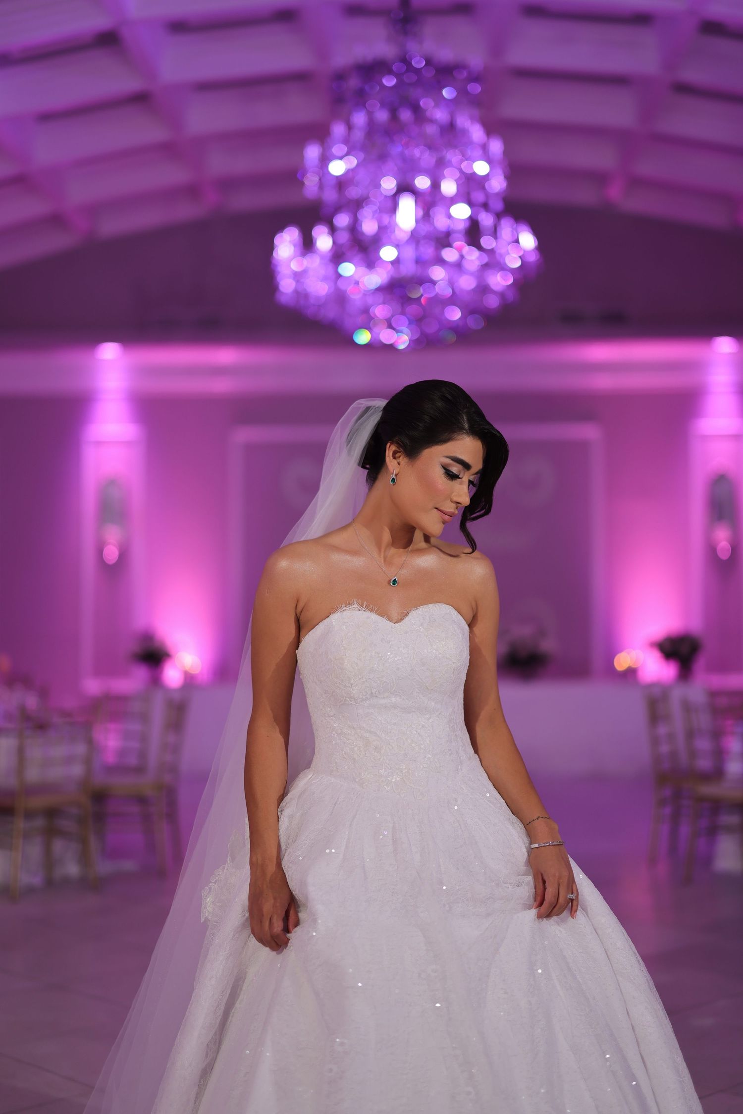 A bride in a sparkly white ballgown stands in an elegant ballroom with purple uplighting and a crystal chandelier.