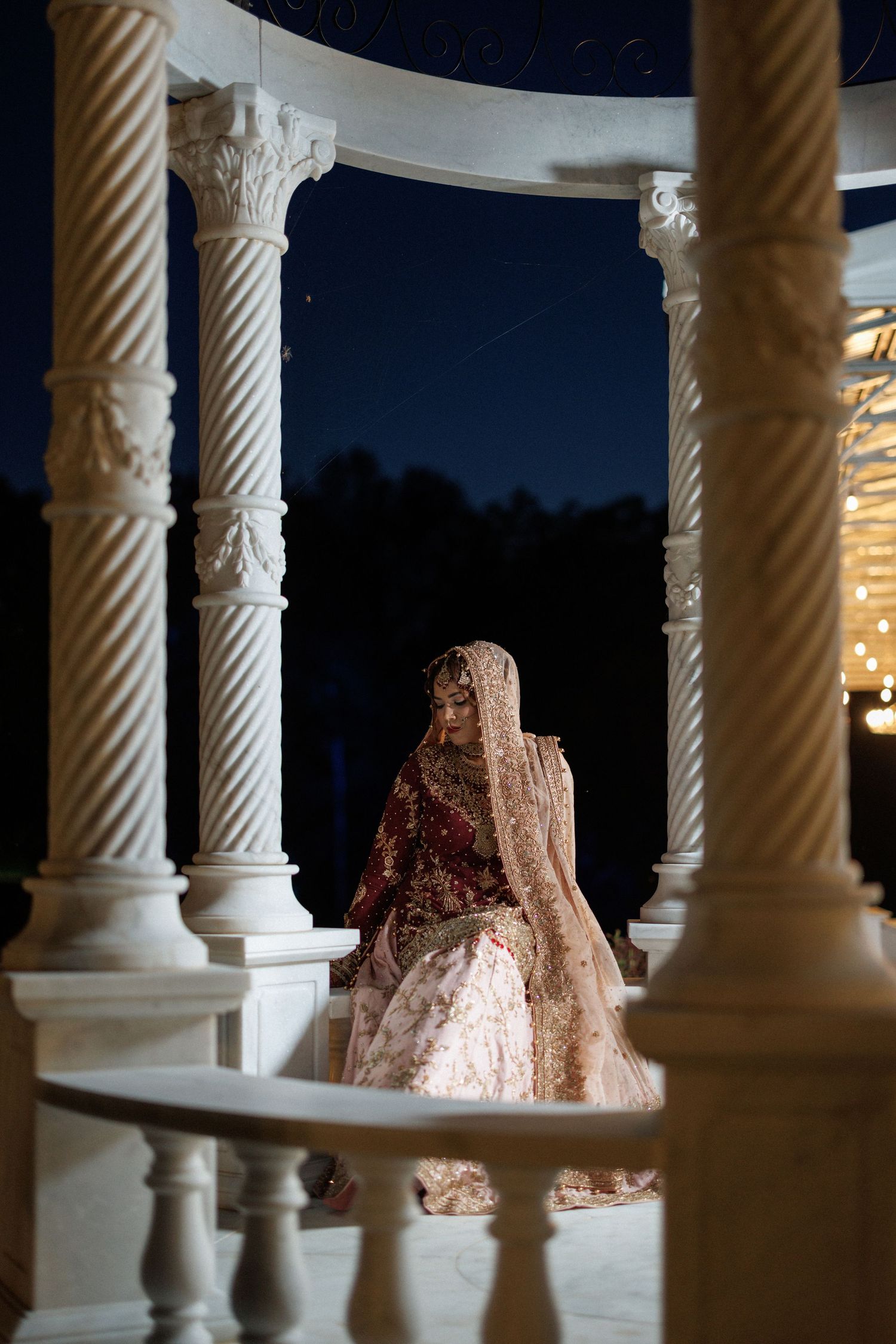 A bride in a sparkly pink and red traditional South Asian lehenga stands between ornate white columns at night.