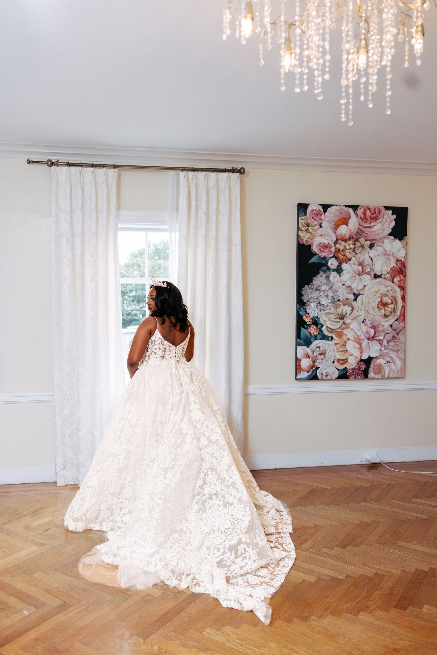 A flowing white lace wedding dress displayed on a hardwood floor with floral artwork and sheer curtains in the background.
