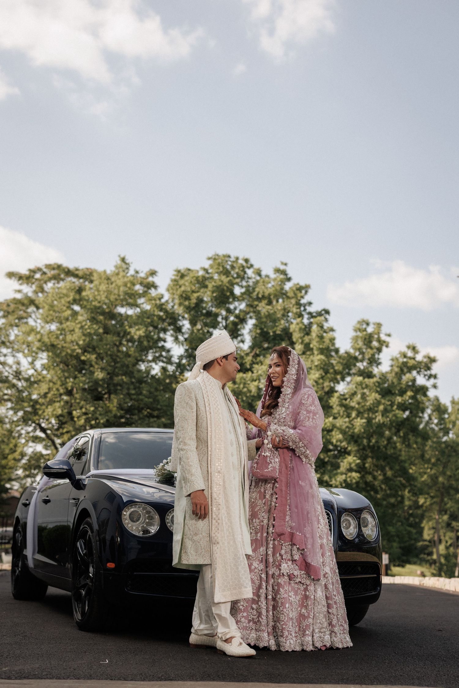 A couple in traditional South Asian wedding attire stands next to a black luxury car during their outdoor celebration.