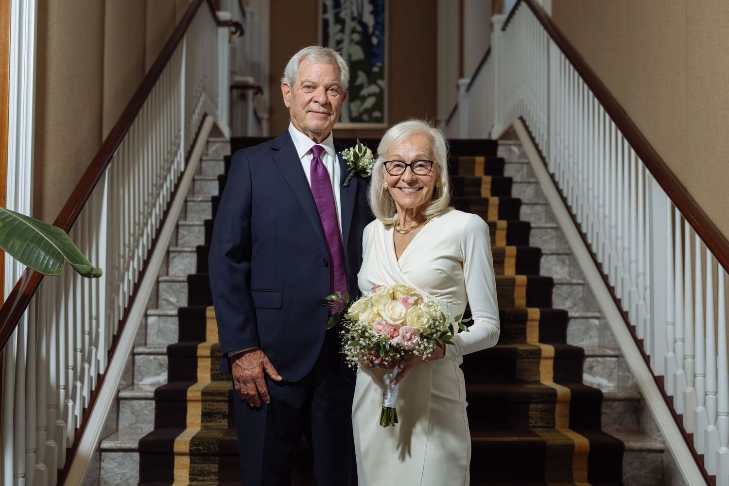 Elegant couple poses on wooden staircase at formal event, with bride holding white floral bouquet.