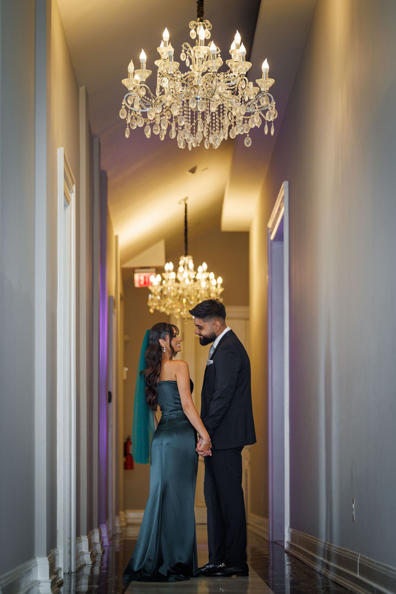 A couple shares an intimate moment in an elegant hallway beneath crystal chandeliers.