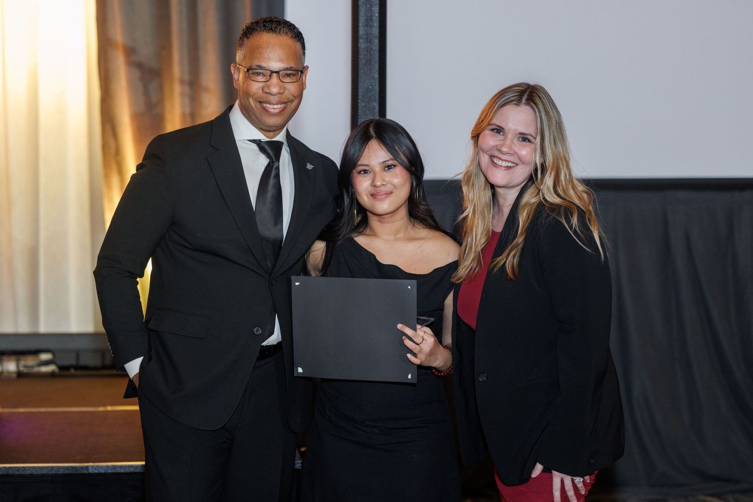 Three professionals posing together at a formal awards ceremony, with woman holding a black certificate folder.