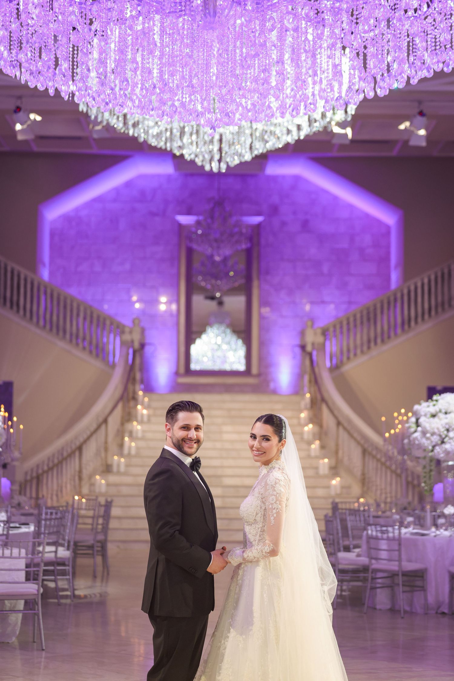 Elegant wedding venue with purple uplighting, crystal chandeliers, and grand white staircases creates a romantic atmosphere.