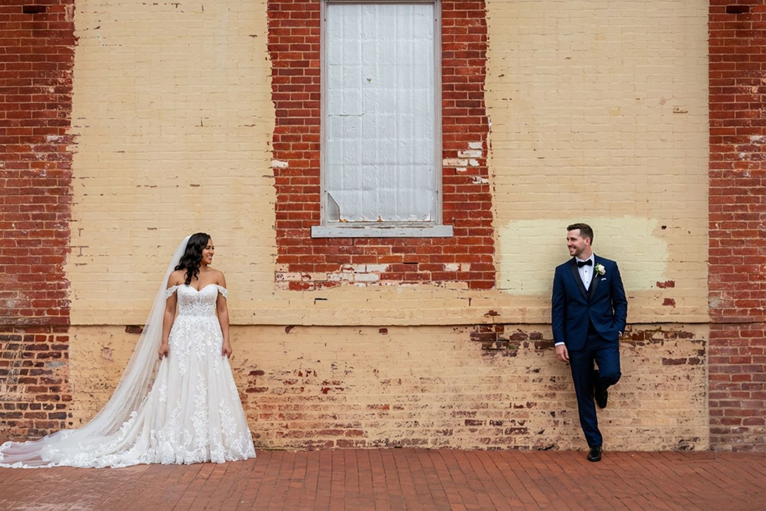 Couple stands in front of rustic brick wall with peeling paint during outdoor wedding photo session.