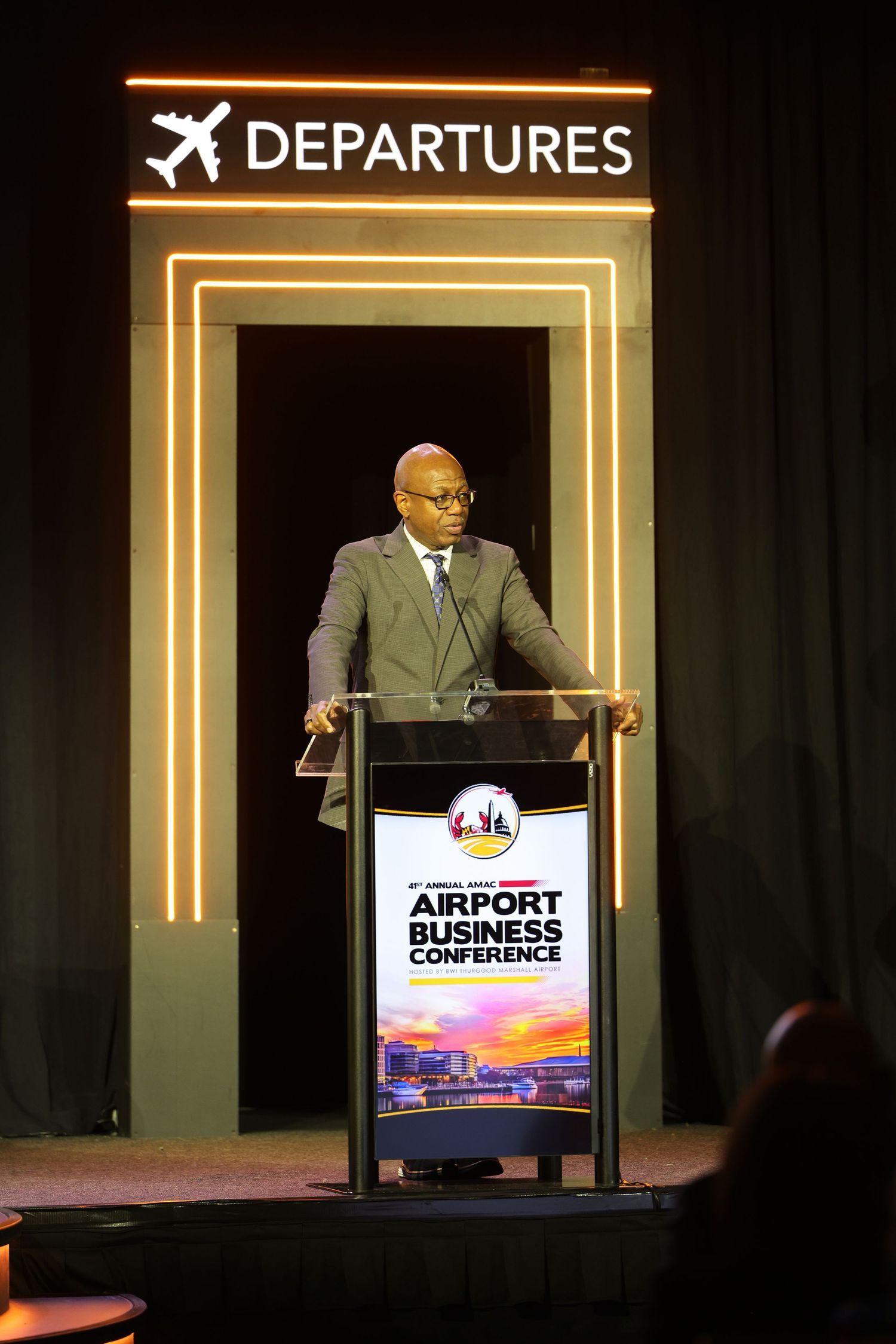 Speaker at Airport Business Conference podium under illuminated Departures sign at aviation industry event.