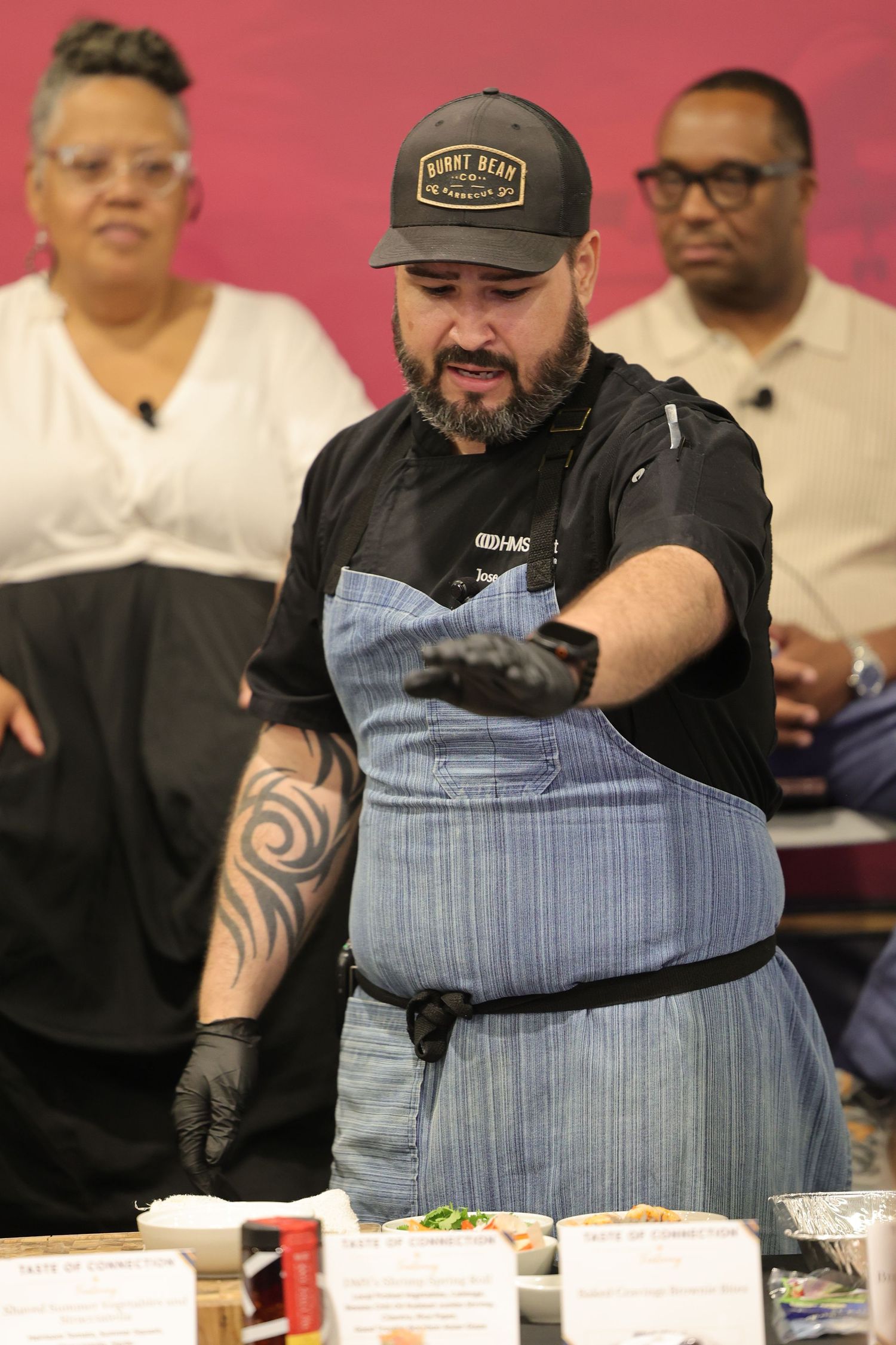 Chef in black cap and apron with tattoos presents food at a cooking demonstration event.