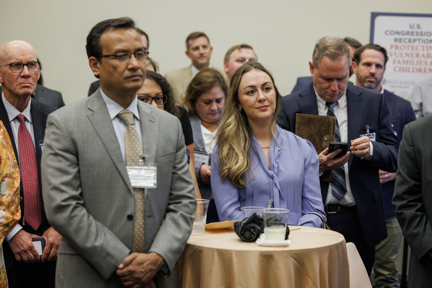 Professionals in business attire stand attentively at a congressional briefing event, with name badges visible.