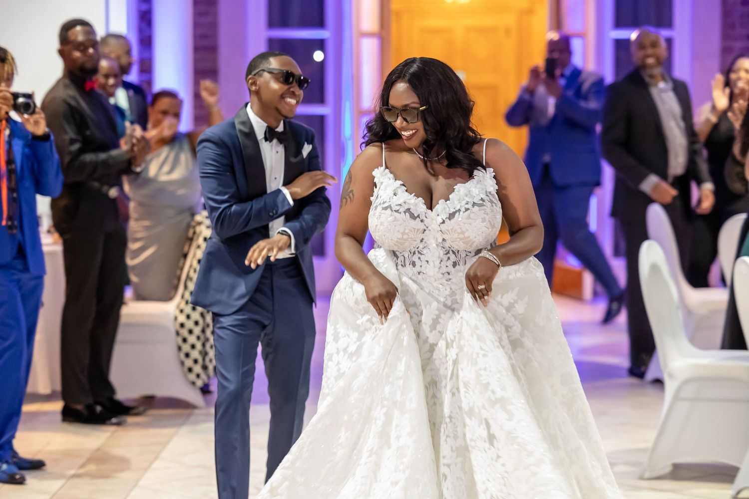 A wedding reception celebration with guests dancing and mingling under purple and orange lighting in an elegant venue.