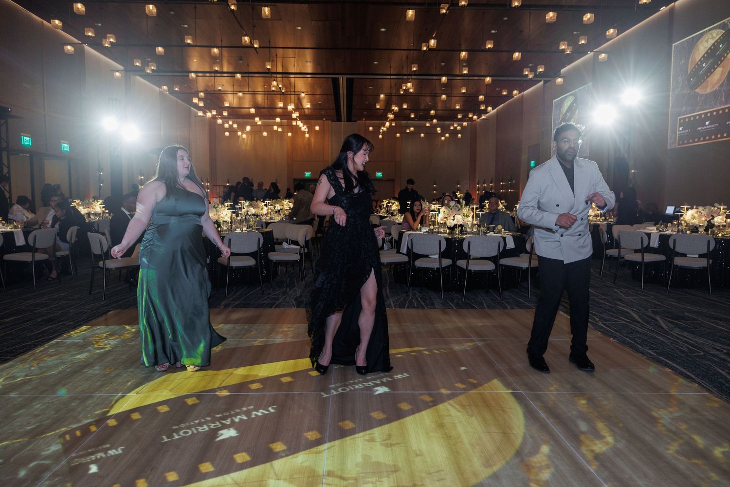 Wedding guests dance on a rustic wooden floor under warm string lights at an indoor reception venue.