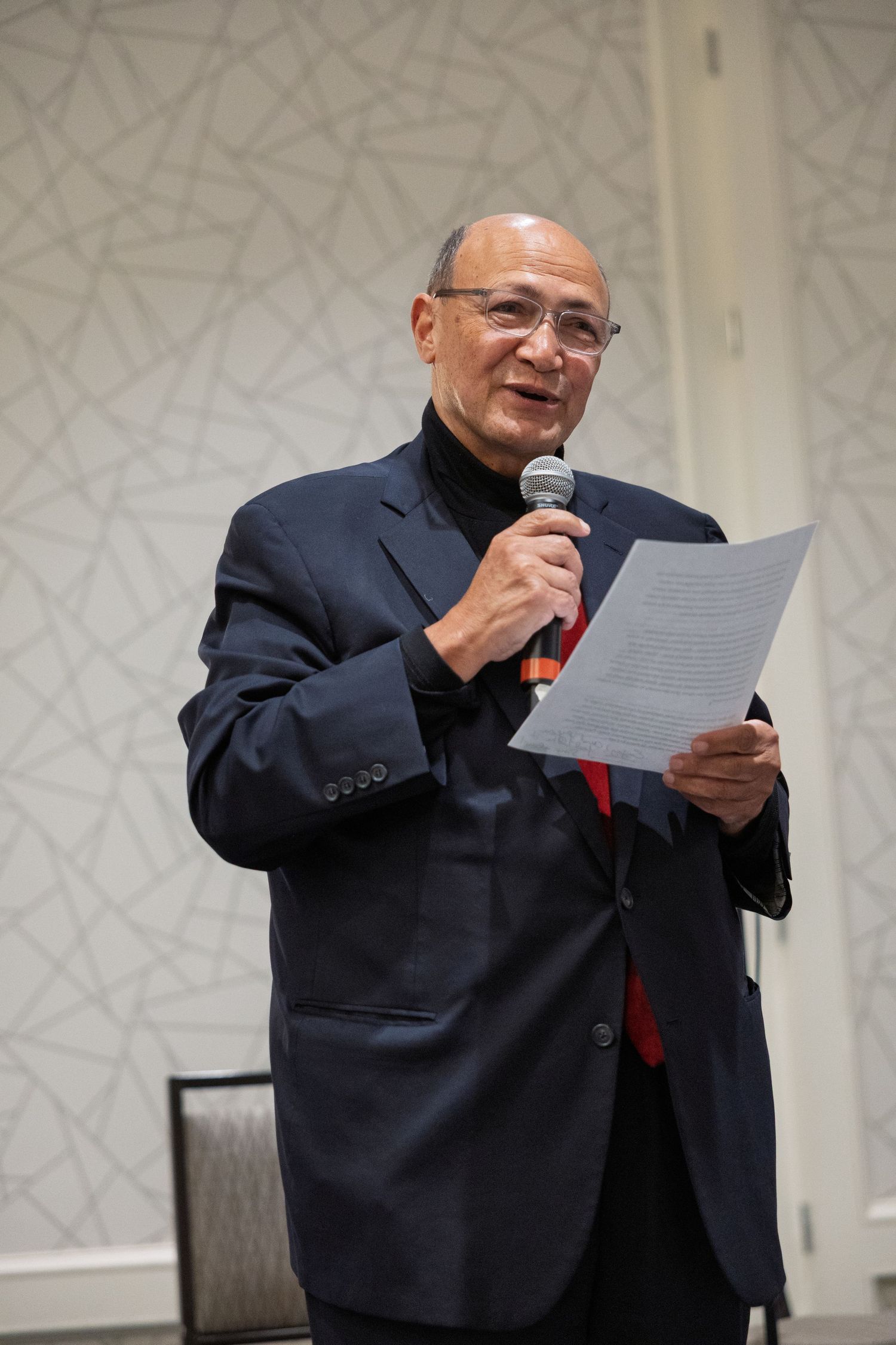 A speaker in a navy suit and red tie reads from papers while holding a microphone during a presentation.