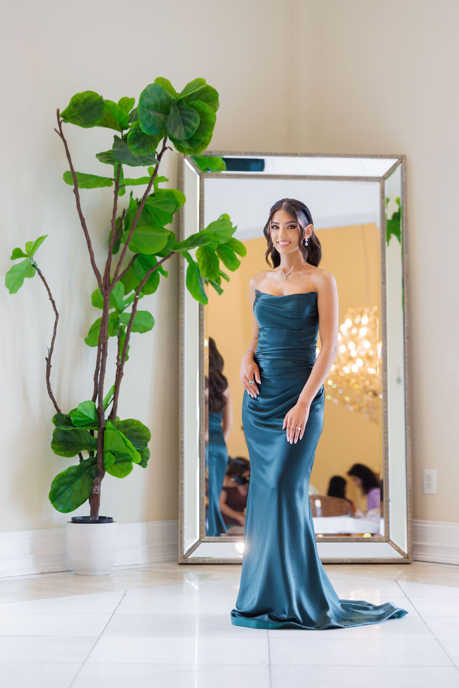 A figure in an elegant teal evening gown poses gracefully in a bright minimalist room with a large fiddle leaf fig plant.