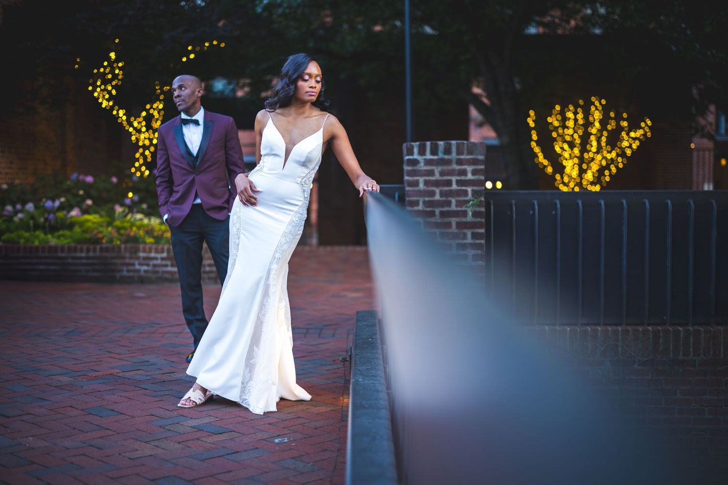 Wedding couple walks together holding hands along a brick pathway at sunset with romantic lighting and string lights.