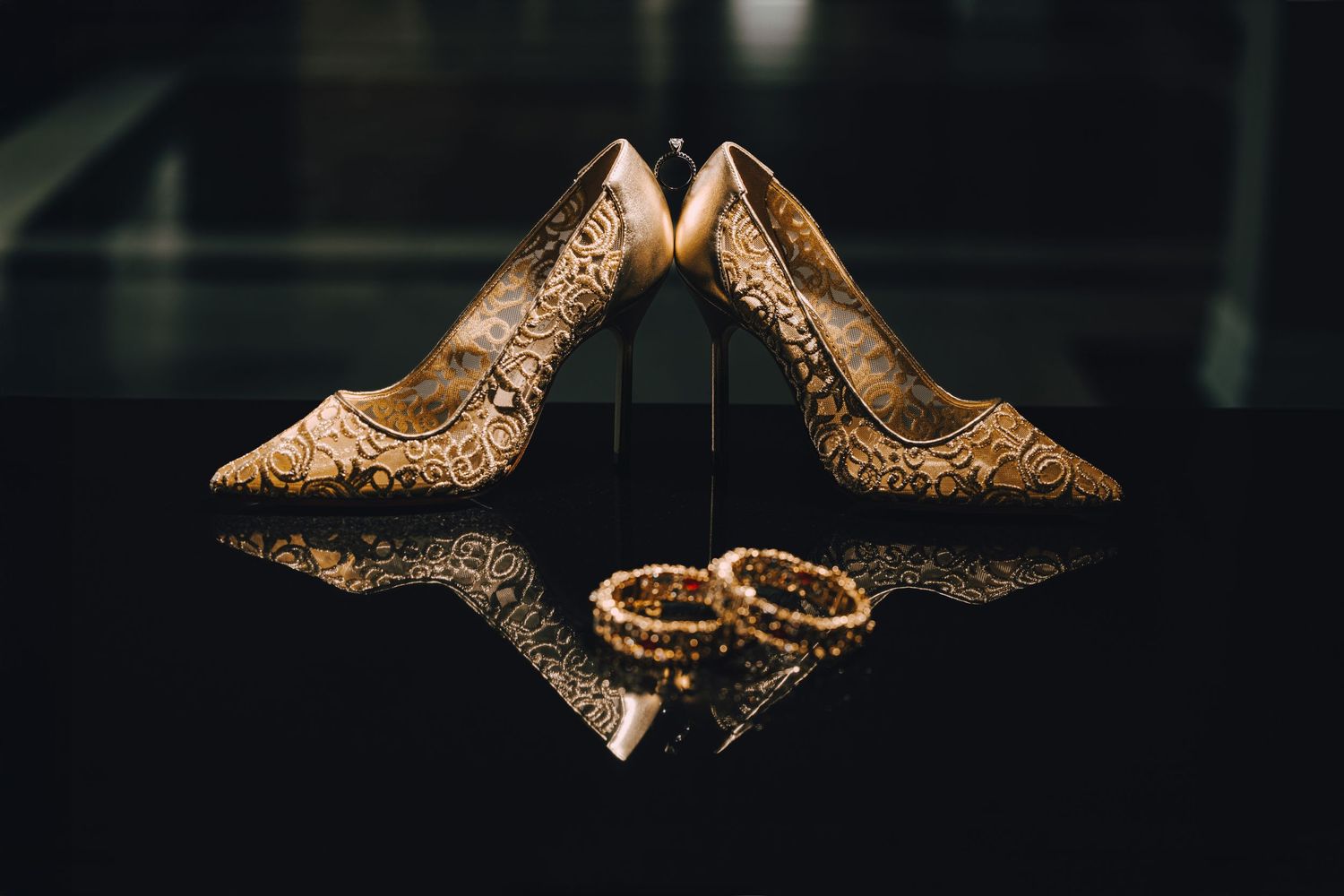 Gold bridal heels and wedding rings styled with reflection on dark surface for wedding photography.