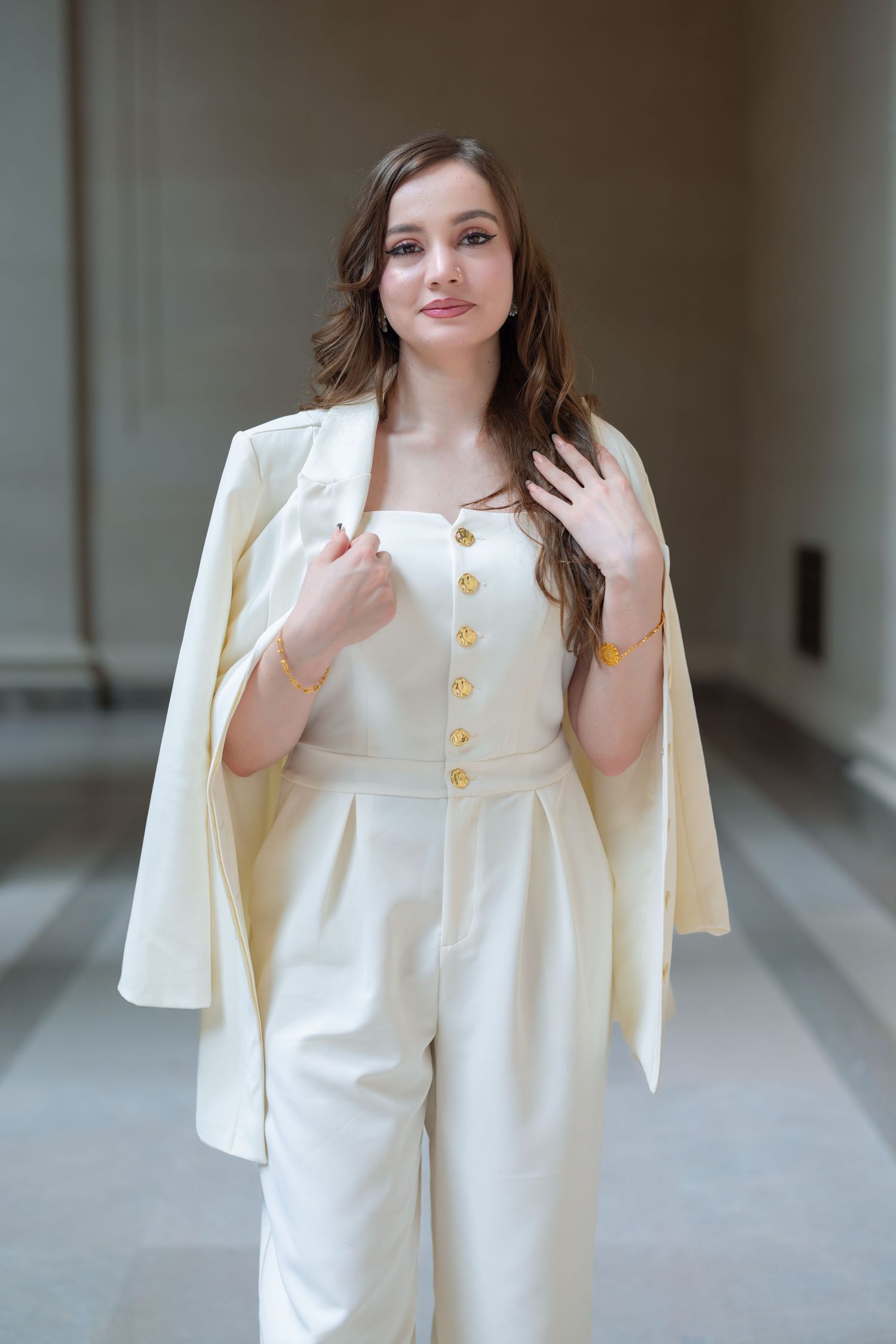 A person wearing a white coordinated outfit with a jumpsuit featuring gold buttons and a matching blazer draped over shoulders.