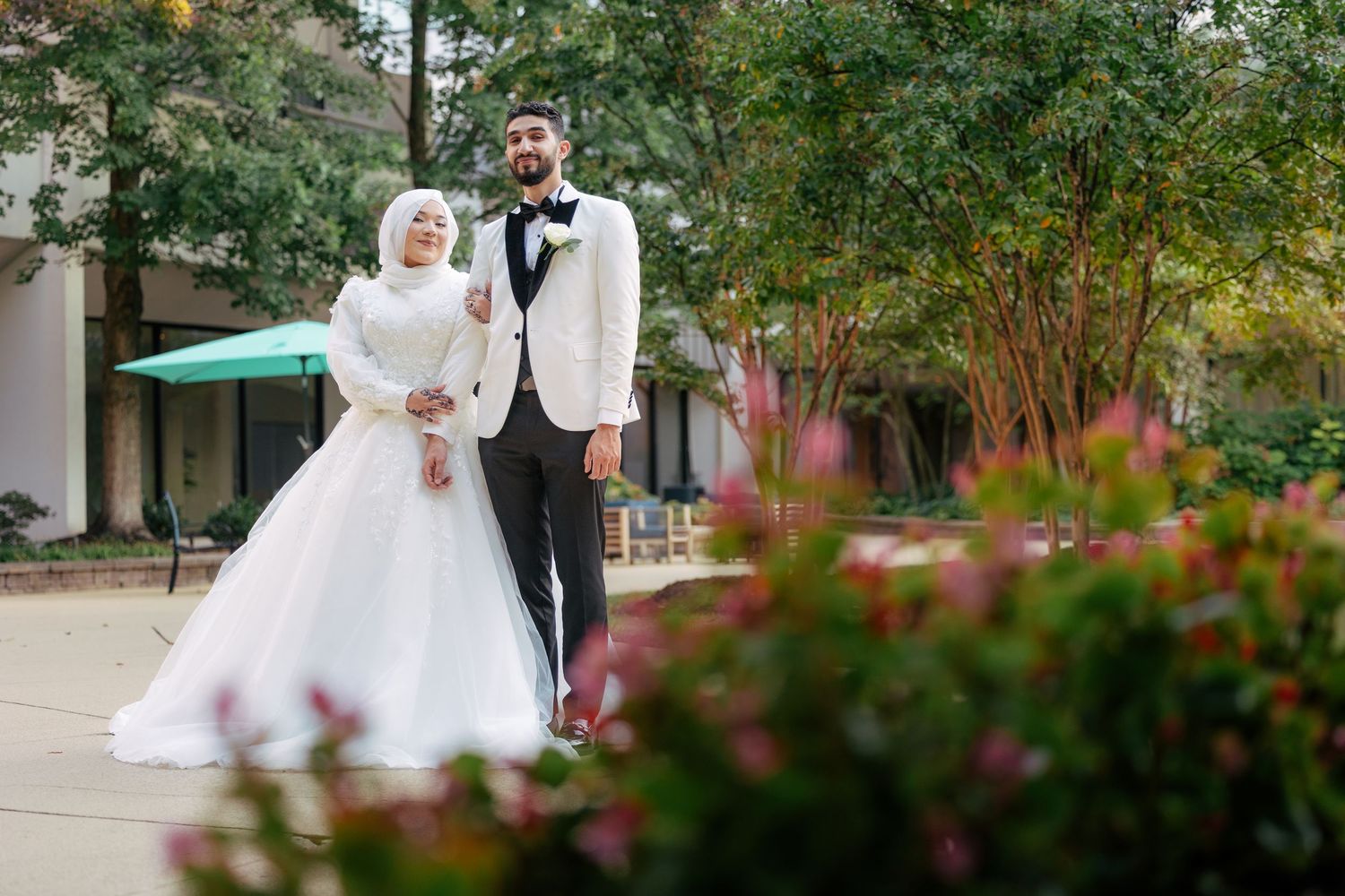 Newlywed couple shares romantic moment in lush garden setting with red flowers in foreground.